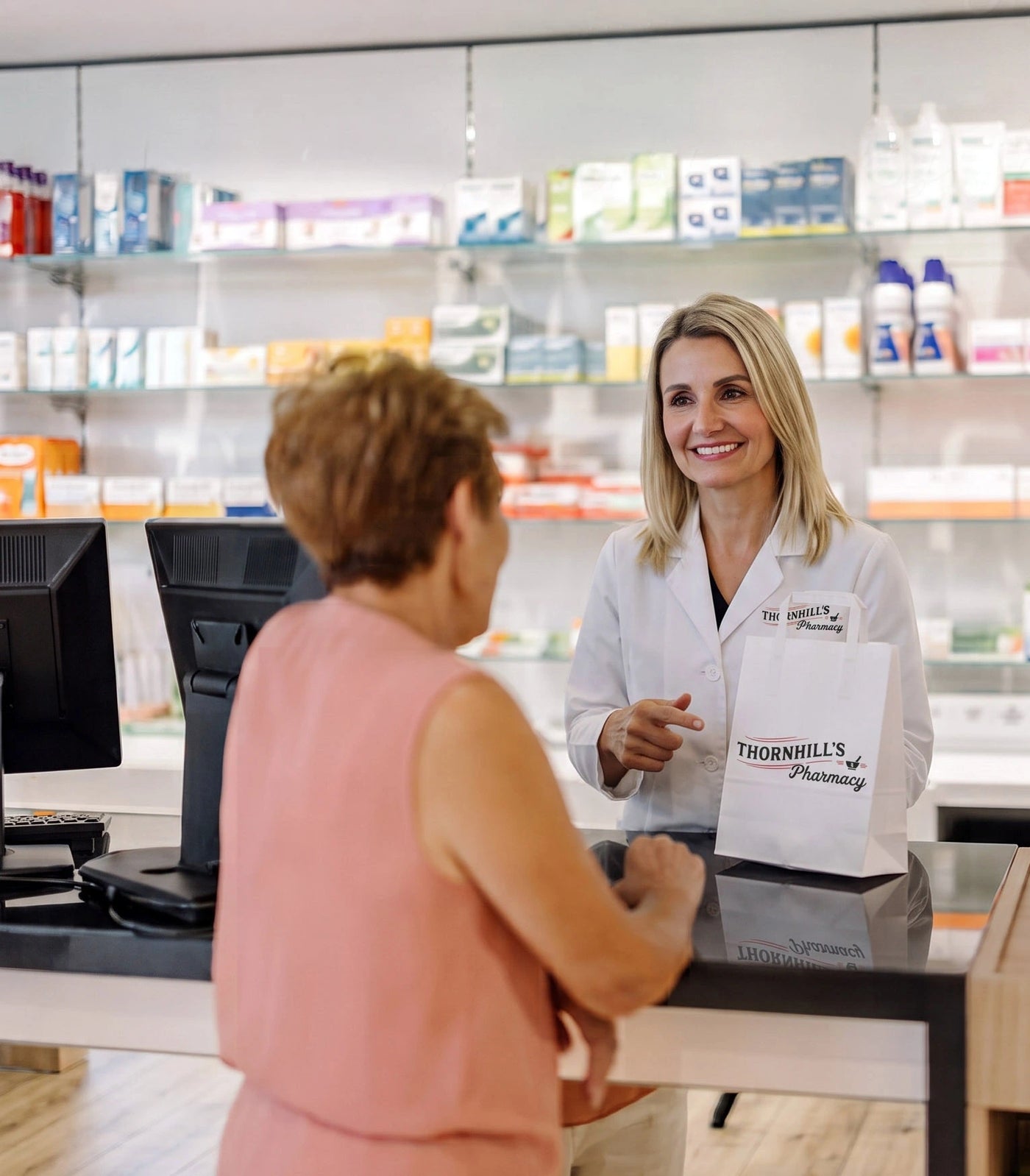 A pharmacist in a white lab coat at Thornhill's Pharmacy speaking with a customer at a pharmacy counter with blurred pharmacy products and items in the background.